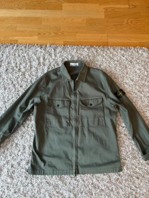Grön overshirt från Stone Island - Snygg grön overshirt från Stone Island med klassisk krage, två bröstfickor med knappar och dragkedja framtill. Ikonisk logotyp på vänster ärm. Perfekt för lager-på-lager och streetwear-stil.