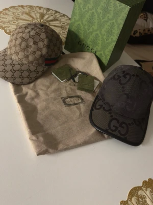 Gucci kepsar med GG-mönster - Två snygga kepsar från Gucci, båda med klassiskt GG-mönster. Den ena är beige med grönt och rött band på sidan, den andra är mörkare med stora GG-loggor. Båda har böjd skärm och justerbar rem bak. Perfekt för dig som vill ha en lyxig och trendig accessoar.