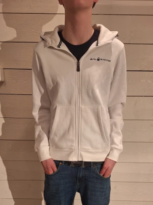 Vit hoodie från Sail Racing - Säljer en clean vit hoodie från Sail Racing med dragkedja framtill och två fickor. Hoodien har logga på bröstet och en liten logga på huvan. Skön passform och mjukt material, perfekt till jeans eller joggers. Modelen är 172 cm!