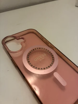 Rosa Guess mobilskal med MagSafe för IPhone 16 - Snyggt rosa mobilskal från Guess för IPhone 16 med genomskinlig design och MagSafe dekorerad med små stenar runt loggan. Skalets material är plast och det har utskärning för kamera och knappar. Perfekt för dig som vill ha både stil och funktion.