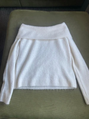 Vit off shoulder stickad tröja - Mysig vit stickad tröja med bred ribbad off shoulder-krage och långa ärmar. Tröjan har en avslappnad passform och ribbade muddar vid ärmslut och nederkant. Perfekt för lager-på-lager och en trendig look.