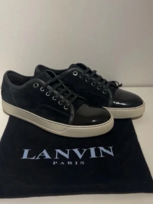 Svarta Lanvins sneakers från Lanvin - Snygga Lanvins svarta sneakers från Lanvin med ovandel i mocka och lackad tå. Skorna har vita sulor, svarta skosnören och metallfärgade öljetter. Klassisk låg modell med exklusiv känsla och diskret logga på påsen. Perfekta för dig som gillar stilrena detaljer.