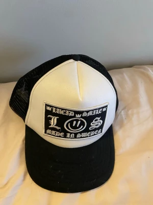Svart vit chrome heart keps - Säljer en svartvit truckerkeps från Chrome hearts med deras legendariska design på framsidan. Kepsen har svart mesh baktill och böjd skärm i svart tyg. Perfekt för dig som gillar streetwear och unik stil.