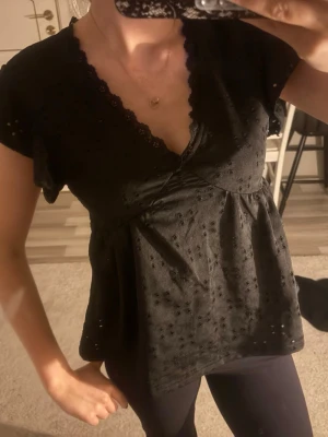 Svart blus med spetsdetaljer - Söt svart blus med v-ringning och spetskant. Blusen har korta ärmar och ett broderat hålmönster över hela tyget. Modellen är lätt utställd och ger en luftig känsla, perfekt till jeans eller kjol.💕