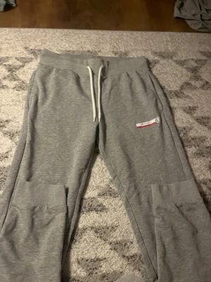 Gråa mjukisbyxor från Champion - Säljer ett par gråa mjukisbyxor från Champion med vita snören i midjan och ribbade muddar vid bensluten. Byxorna har en liten Champion-logga på ena benet och är perfekta för chill dagar. Klassisk passform och mjukt material.