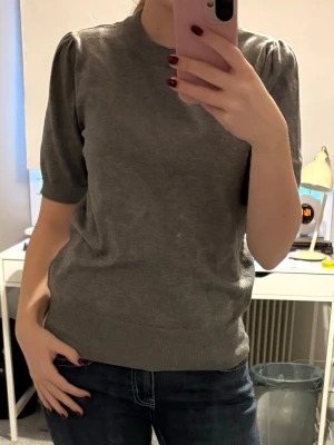 Grå topp med puffärm - Snygg grå topp med puffärm och rund hals. Modellen är något loose fit och har ribbade muddar vid ärmslut och nederkant. Perfekt till jeans eller kjol för en clean look. SPEGLN ÄR LITE SMUTSIG