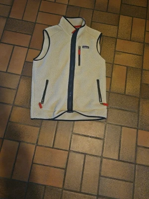 Beige fleeceväst från Patagonia - Säljer en beige fleeceväst från Patagonia med svarta detaljer och röda dragkedjor. Västen har hög krage, två fickor framtill och hel dragkedja. Perfekt för lager-på-lager och outdoor-vibe.