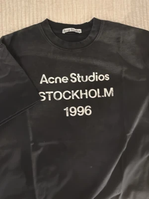 Svart Acne Studios t-shirt - Säljer en svart t-shirt från Acne Studios med vit text 'Acne Studios STOCKHOLM 1996' på bröstet. Klassisk passform och rund halsringning. Perfekt för dig som gillar stilrena och ikoniska plagg.