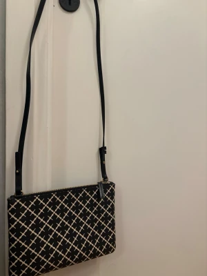 Svart axelväska med rutigt mönster - Snygg svart axelväska med vitt rutigt mönster och justerbar axelrem.!!!som går att ta av!!!Väskan har dragkedja upptill och är tillverkad i ett tåligt syntetmaterial. Perfekt för dig som vill ha en stilren och trendig accessoar till din outfit.
