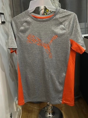 Grå och orange träningströja Puma - Sportig t-shirt från Puma i grått med orangea sidopaneler och stort orange logotryck på bröstet. Tillverkad i ett lätt och snabbtorkande material som passar perfekt till träning eller gym. Klassisk rund hals och korta ärmar. Har puma rivningar som modell.