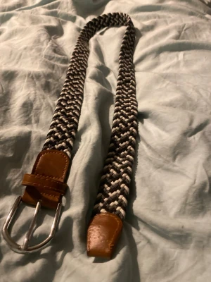 Svartvitt flätat bälte med läderdetaljer - Snyggt svartvitt flätat bälte med bruna läderdetaljer vid spännet och änden. Bältet har ett klassiskt metallspänne och är gjort i ett elastiskt material som ger en bekväm passform. Perfekt för att lyfta vilken outfit som helst.