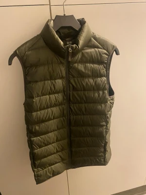 Olivgrön dunväst från Uniqlo - Snygg olivgrön dunväst från Uniqlo med hög krage och dragkedja framtill. Västen har en quiltad design och är lätt vadderad, perfekt för lager-på-lager. Passar till både chill och streetwear-stil.