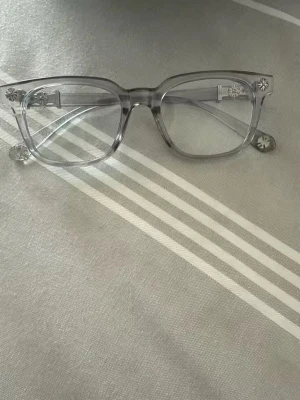 Chrome hearts fyrkantiga glasögon - Säljer ett par stilrena chrome hearts glasögon med transparent, fyrkantig båge i plast. Bågarna har diskreta blommönster på sidorna för en unik touch. Perfekta för dig som gillar en modern och clean look.