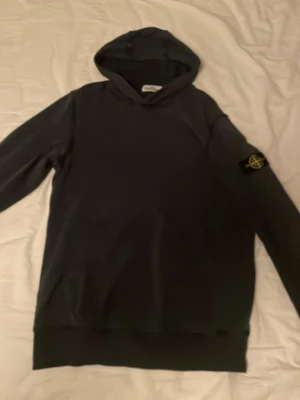 Mörk blå hoodie från Stone Island - Säljer en mörk blå hoodie från Stone Island med den klassiska loggan på ärmen. Hoodien har huva och ribbade muddar vid ärmslut och nederkant. Tillverkad i mjukt bomullsmaterial som är skönt att bära. Perfekt för dig som gillar streetwear och vill ha en clean look.