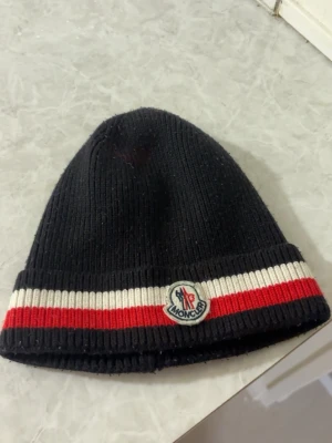 Moncler mössa  - En svart moncler mössa med vit och röda ränder. Har lite missfärgning som syns på första bilden men är ej så tydlig. Säljer den billigt pga att den inte används längre samt den lilla otydliga missfärgningen
