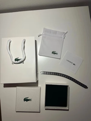 Lacoste silverfärgat armband - Säljer ett stilrent armband från Lacoste i silverfärgad metall med klassiska krokodilloggan ingraverad på länken. Armbandet har ett rutigt mönster på varje länk och kommer med original ask, påse och certifikat. Perfekt accessoar för dig som gillar clean design.