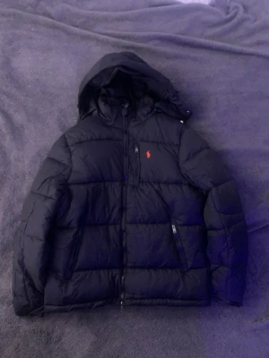 Svart pufferjacka från Polo Ralph Lauren - Svart pufferjacka från Polo Ralph Lauren med röd broderad logga på bröstet. Jackan har huva, dragkedja framtill och två sidofickor med dragkedja. Perfekt för kalla dagar och har en klassisk, sportig look.