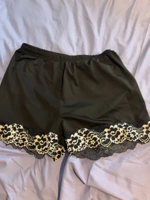 Svarta shorts med spetskant - Snygga svarta loungewear-shorts med elastisk midja och dekorativ spetskant i beige och svart längs bensluten. Perfekta för chill hemma eller mysiga kvällar. Mjukt och lätt material som känns skönt mot huden.