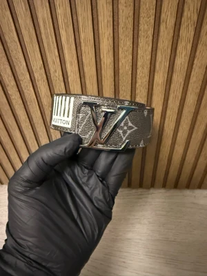 Bälte - Snyggt bälte. Bältet har detaljer i silver och vitt samt en unik etikett med ränder och Vuitton-tryck. Perfekt accessoar för att lyfta din outfit.