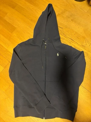 Mörkblå hoodie från Polo Ralph Lauren - Snygg mörkblå hoodie från Polo Ralph Lauren med dragkedja framtill och klassisk vit logga på bröstet. Hoodien har justerbar huva med snören och två fickor framtill. Tillverkad i mjukt bomullsmaterial som känns skönt mot huden.