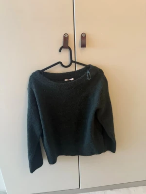 Mörkgrön stickad tröja - Mysig mörkgrön stickad tröja med rund halsringning och långa ärmar. Tröjan har en enkel, rak passform och är gjord i ett fluffigt, mjukt material som känns skönt mot huden. Perfekt för kyliga dagar och enkel att matcha med olika outfits.