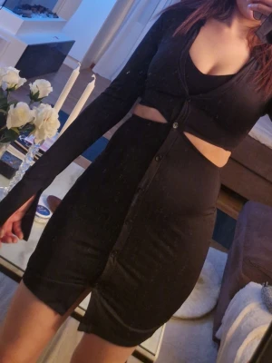 Svart kort klänning med cut out - Trendig svart kort klänning med cut out-detalj i midjan och knappar framtill. Klänningen har lång ärm och en kroppsnära passform som framhäver siluetten. Perfekt för dig som vill ha en edgy och modern look.