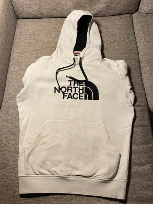 Vit hoodie från The North Face - Säljer en vit hoodie från The North Face med stor svart logga på bröstet. Hoodien har en klassisk känguruficka, dragsko i huvan och mjukt bomullsmaterial. Insidan av huvan är svart för en snygg kontrast. Perfekt för dig som gillar streetwear och enkel stil. Pris kan diskuteras.