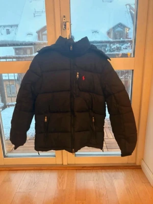Svart dunjacka från Ralph Lauren - Svart puffer dunjacka från Ralph Lauren med huva och dragkedja framtill. Jackan har två fickor med dragkedja och en liten röd logga på bröstet. Perfekt för kalla vinterdagar och riktigt skön att bära.
