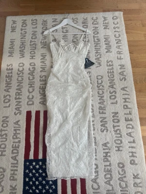 Vit spetsklänning med snörning - Elegant långklänning i vit spets med tunna axelband och snörning framtill. Klänningen har ett blommigt mönster och figurnära passform som ger en romantisk look. Perfekt för sommarens speciella tillfällen.