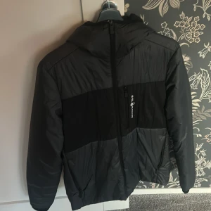 Svart Sail Racing pufferjacka M - Säljer en svart pufferjacka från Sail Racing i storlek medium. Jackan har huva, dragkedja framtill och en bröstficka med logga. Materialet är nylon och polyester med Primaloft-foder som håller dig varm. Snygg och sportig look med diskreta detaljer.