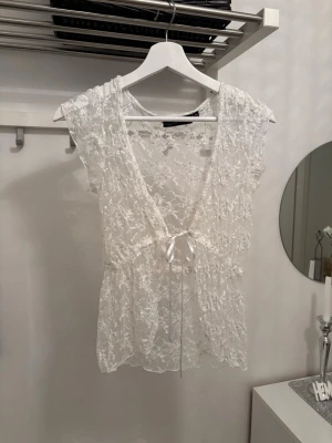Vit spetsblus med knyt framtill - Superfin vit blus i spets med v-ringning och knyt framtill. Blusen är ärmlös och har ett romantiskt, transparent utseende med blommiga spetsdetaljer. Perfekt att styla lager på lager eller som den är för en trendig look.