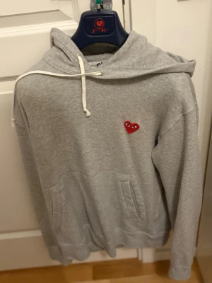 Grå hoodie Comme des Garçons Play - Säljer en grå hoodie från Comme des Garçons Play med det ikoniska röda hjärtat med ögon broderat på bröstet. Hoodien har huva med vita snören och en stor magficka. Tillverkad i mjukt bomullsmaterial, perfekt för en chill och stilren look. Varan ör i princip nyskick.