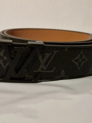Louis Vuitton monogram bälte svart - Snyggt svart bälte från Louis Vuitton med klassiskt monogrammönster och stor LV-logga som spänne. Bältet har en slät, brun insida och är gjort i läder med exklusiv känsla. Perfekt accessoar för att lyfta vilken outfit som helst.