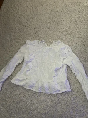 Vit volangblus från Kappahl - Söt vit blus från Kappahl med volangdetaljer framtill och längs ärmarna. Blusen har rund halsringning och är tillverkad i ett lätt, strukturerat tyg som ger en luftig känsla. Perfekt för dig som gillar romantisk och trendig stil.