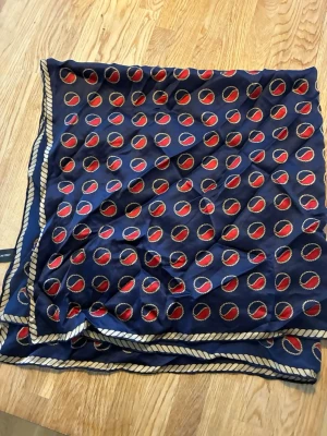 Busnel Mörkblå sidenscarf med repmönster - Snygg mörkblå scarf i 100% siden med repmönstrad kant och röda cirklar med repdetaljer. Perfekt accessoar för att lyfta din outfit med en klassisk och färgstark touch. Fyrkantig form och mjukt, glansigt material.