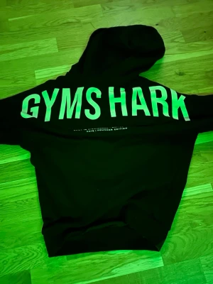 Svart hoodie från Gymshark - Säljer en svart hoodie från Gymshark med stor vit logga över ryggen. Hoodien har en klassisk huva och är perfekt för dig som gillar streetwear eller träningskläder. Materialet är mjukt och bekvämt, perfekt för chill dagar eller gymmet.