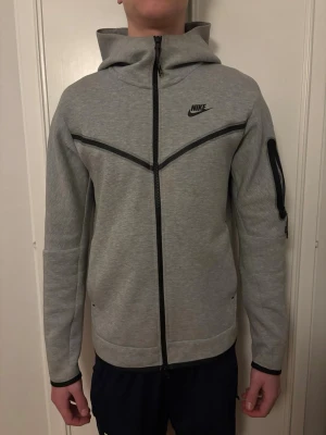 Grå Nike Tech Fleece hoodie - Snygg grå Nike Tech Fleece hoodie med hel dragkedja och svarta detaljer. Har huva, två fickor fram och en extra ficka med dragkedja på ärmen. Nike-logga på bröstet. Tillverkad i mjukt och bekvämt material.
