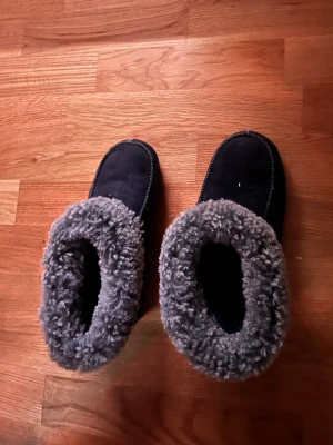 Mörkblå tofflor med grått foder - Mysiga mörkblå tofflor med fluffigt grått foder runt öppningen. Tofflorna har en enkel design med rund tå och är perfekta för att hålla fötterna varma hemma. Materialet ser ut att vara syntet eller fleece för extra komfort.