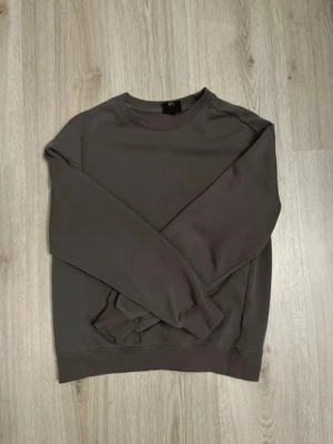 Mörkbrun sweatshirt med rund hals - En stilren mörkbrun sweatshirt med rund halsringning och långa ärmar. Tröjan har ribbade muddar vid ärmslut och nederkant, perfekt för en avslappnad och trendig look. Passar bra till jeans eller mjukisbyxor.