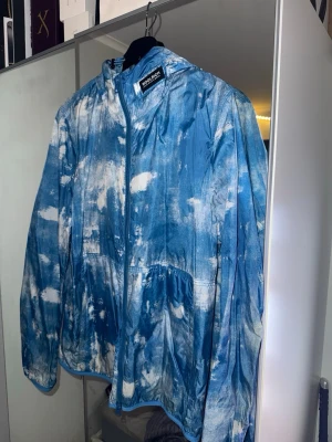 Blåvit vindjacka från Woolrich - Säljer en blåvit vindjacka från Woolrich med unikt batikmönster. Jackan har dragkedja framtill, hög krage och två sidofickor. Perfekt för dig som vill sticka ut med en färgstark och lätt jacka. Passar till både vår och sommar.