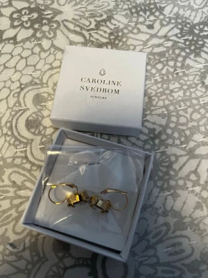 Caroline Svedbom örhängen - Säljer ett par örhängen från Caroline Svedbom i färgen Golden Shadow. Örhängena har gnistrande ljusgula kristaller och guldfärgade detaljer. Perfekta för att ge din look lite extra bling!