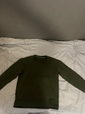 Mörkgrön sweatshirt från Sail racing - Säljer en mörkgrön sweatshirt från Sail racing med rund halsringning och diskret logga på bröstet. Tröjan har långa ärmar och ribbade muddar vid ärmslut och nederkant. Perfekt för en avslappnad och trendig look.