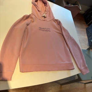 Rosa hoodie från GANT med tryck - Säljer en snygg rosa hoodie från GANT med vit snörning i huvan och tryckt logga på bröstet. Tröjan har lång ärm, ribbade muddar och är gjord i mjuk bomull. Perfekt för chill dagar eller när du vill ha en clean och stilren look. Mycket bra skick, se muddar