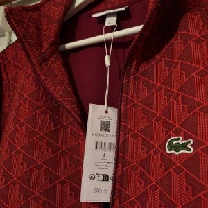 Röd mönstrad ziptröja från Lacoste - Röd ziptröja från Lacoste, köptes i Cypern för 235 euro vilket i kronor är ca 2600kr. Säljer för att tycker jag är lite för stor för tröjan annars jätte nöjd med koftan