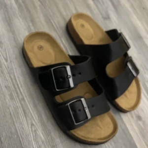 Svarta sandaler med spännen - Snygga svarta sandaler med två breda remmar och stora silverspännen. Fotbädden är beige och ergonomiskt formad för extra komfort. Perfekta för varma dagar och enkel att matcha med olika outfits.