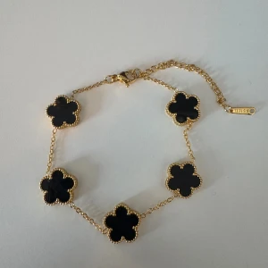 Guldarmband med svarta blommor - Snyggt armband i guld med fem svarta blommor som detaljer. Kedjan är tunn och elegant, och blommorna har en fin guldkant som ger en lyxig känsla. Perfekt accessoar för att lyfta din outfit med en trendig touch.