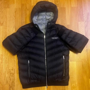 Svart dunjacka från Armani Exchange - Säljer en svart dunjacka från Armani Exchange med huva, äkta dun, broderi och silverfärgade dragkedjor. Jackan har två framfickor med dragkedjor och är perfekt för kyligare väder. Storlek M