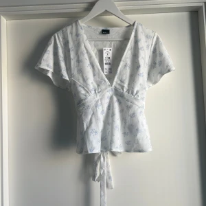Vit blommig omlottopp från Gina Tricot - Supersöt vit omlottopp från Gina Tricot med ljusblått blommönster. Toppen har djup v-ringning, korta ärmar och knytband i midjan för en snygg passform. Perfekt för vår och sommar med sin lätta bomullskänsla och feminina look.