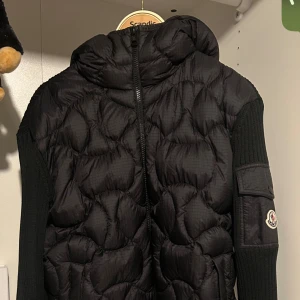 Svart dunväst från Moncler - Svart dunväst från Moncler med quiltad framsida, ribbstickade sidor och ärmar samt huva. Västen har dragkedja framtill och en ficka med Moncler-logga på ärmen. Perfekt för lager-på-lager och snygg streetstil.