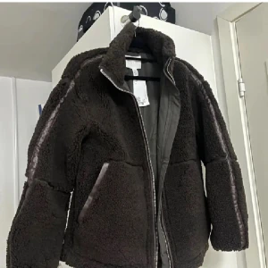 Mörkbrun teddyjacka med dragkedja - Mysig mörkbrun teddyjacka från H&M med hög krage och hel dragkedja framtill. Jackan har detaljer i skinnimitation längs ärmar och vid fickor, samt en schysst loose passform. Perfekt för kalla dagar när du vill vara både varm och snygg. Köpt här på plock, prislapp kvar. Passar mig som vanligtvis har 32/xs